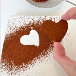 Cœur en chocolat tenu entre les doigts au-dessus d’une assiette saupoudrée de cacao